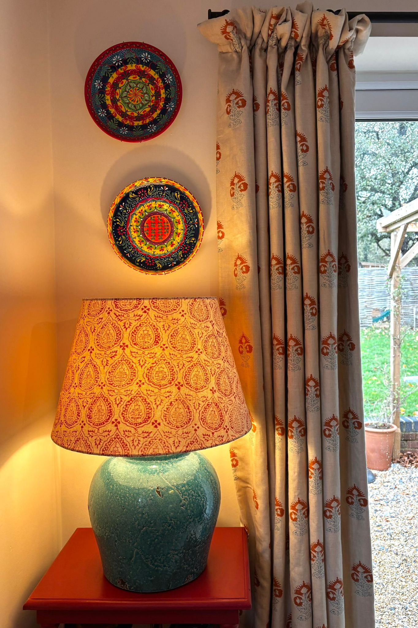 Interior vignette with hand-painted decorative wall plates, paisley lampshade on a blue ceramic lamp and patterned curtains opening to a garden