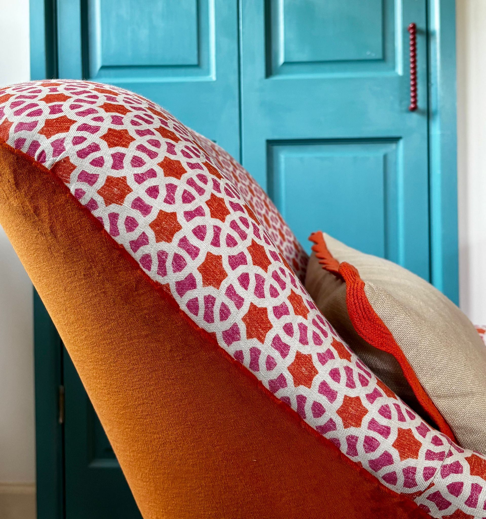 Close-up of an orange velvet armchair with a pink and orange geometric block-print cushion against a teal blue panelled door