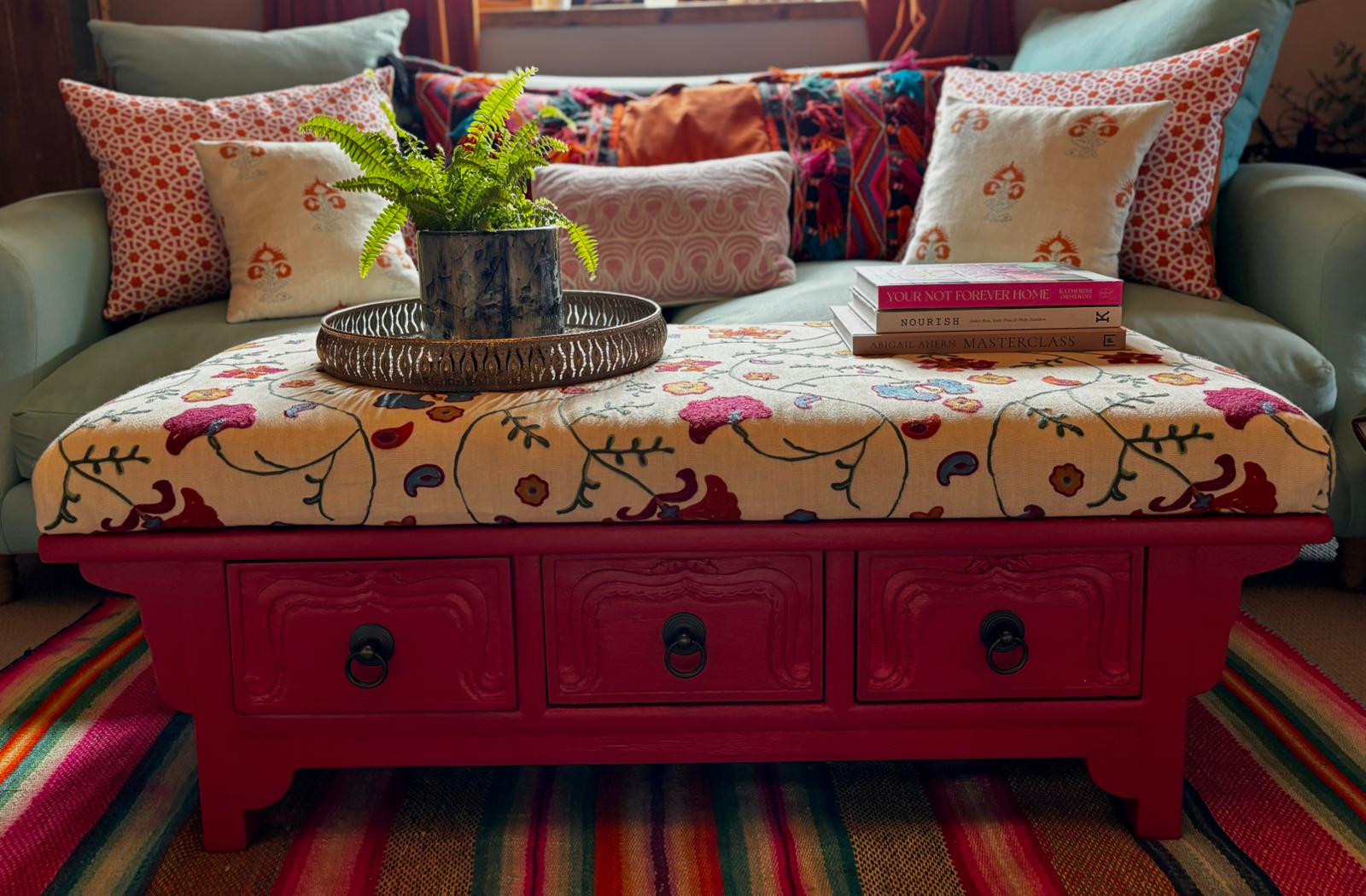 Pink lacquered coffee table topped with a suzani-embroidered cushion, set against a sage sofa layered with block-print cushions and books